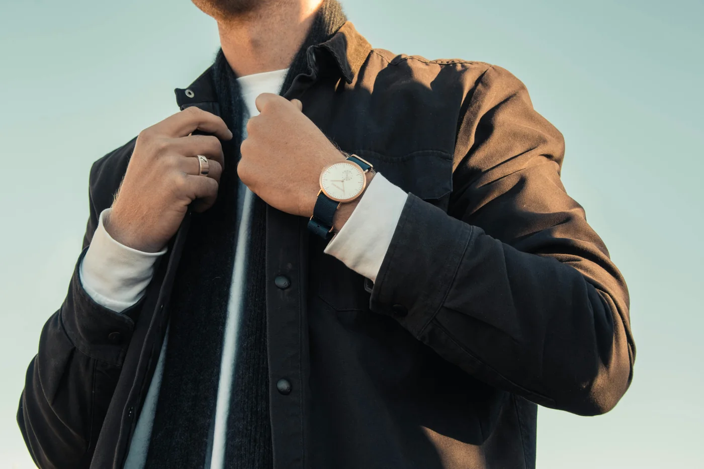 Homme portant une veste à col chemise noire ouverte sur un pull et un tee-shirt blanc, une montre au poignet